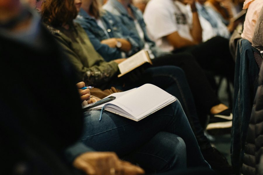 Blick in eine Reihe an Konferenzteilnehmenden. Es sind die Oberkörper und übereinandergeschlagenen Beine der sitzenden Menschen zu sehen. Auf den Beinen zum Teil Notizbücher.